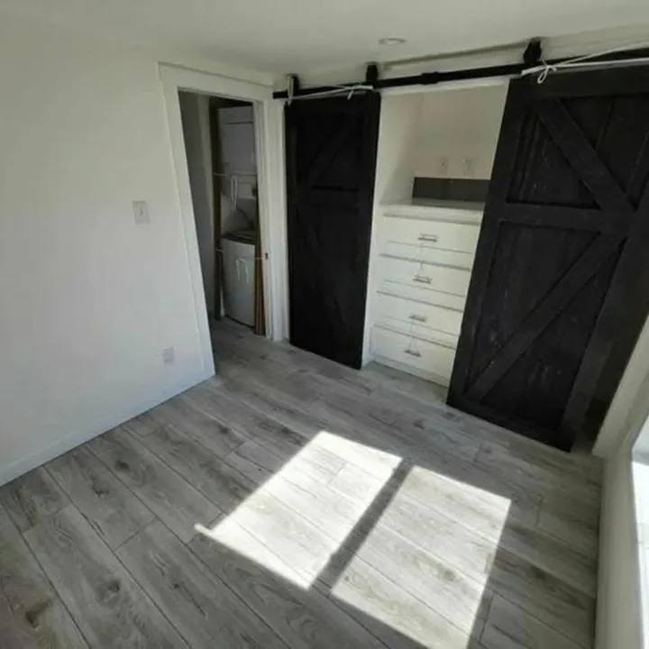 Modern luxury tiny home interior featuring a bright open-concept living area with light grey wood-look flooring and crisp white walls, showcasing a space-saving bedroom with a built-in white dresser and a dual black sliding barn door closet system, alongside a glimpse into a functional laundry nook for a sophisticated move-in ready turn-key residential design.