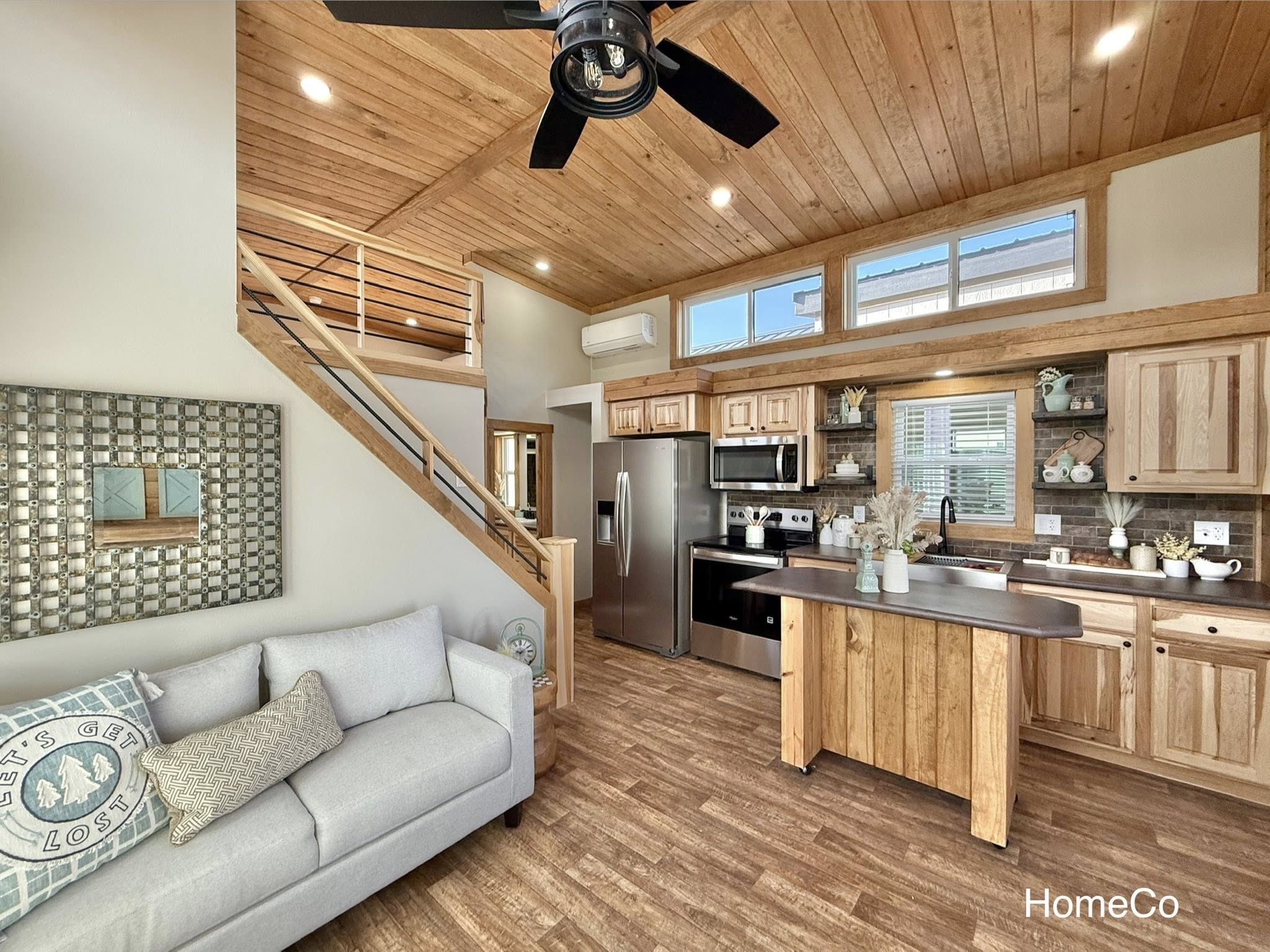 Cozy living space with a high wooden ceiling, modern kitchen, lofted area, light gray sofa, decorative pillows, rustic accents, and large windows.