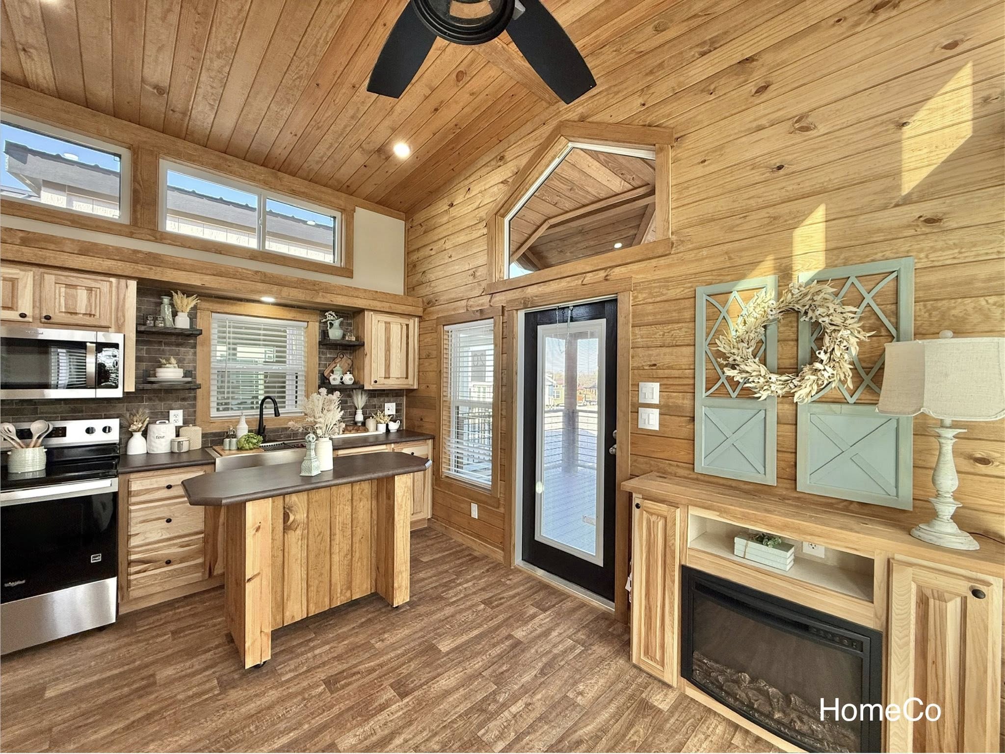 Cozy cabin interior with wooden walls and ceiling, featuring a small modern kitchen, island, and decorative elements. Sunlight streams through high windows.