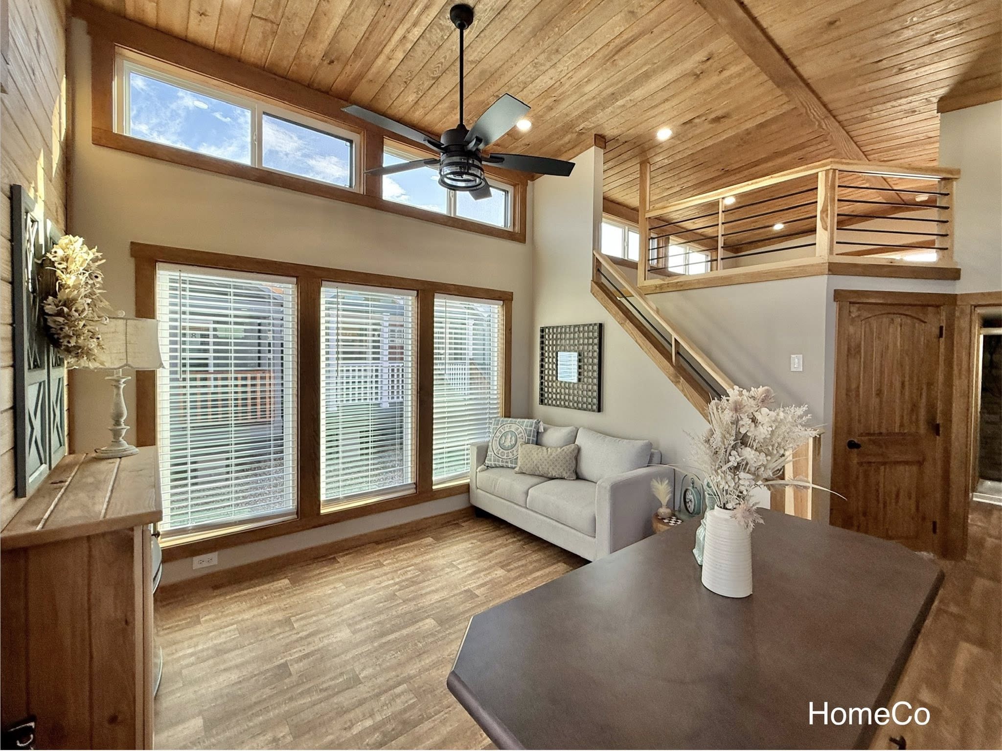 Cozy living room with warm wood accents, large windows, a gray couch, decorative pillows, a table with a vase, and a ceiling fan. Upper loft area in view.