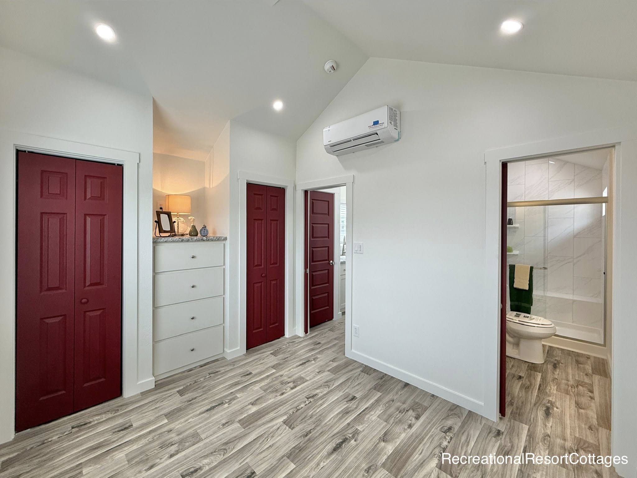 Compact room with red doors, a light wood floor, and white walls. Features a mini-split AC, a mirrored cabinet, and a bathroom with modern fixtures.