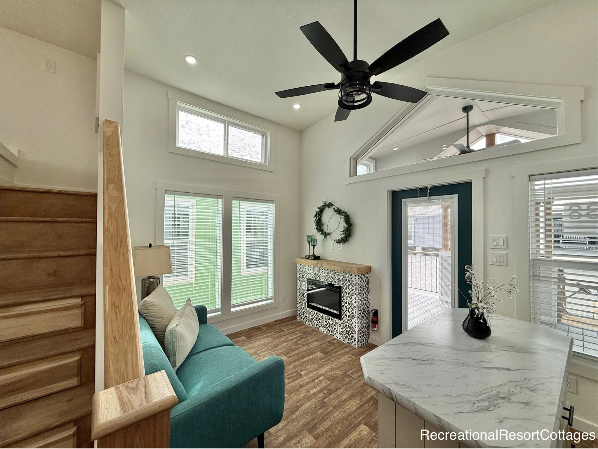 Bright, modern living room with high ceiling, natural light. Features a teal sofa, patterned fireplace, marble counter, and decorative wreath. Cozy ambiance.