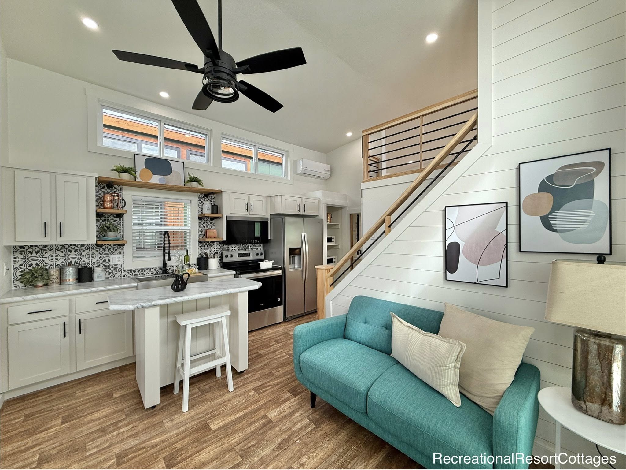 Modern tiny home interior with a teal sofa, abstract art, and white shiplap walls. Bright kitchen with stainless steel appliances and bar seating. Cozy and inviting.