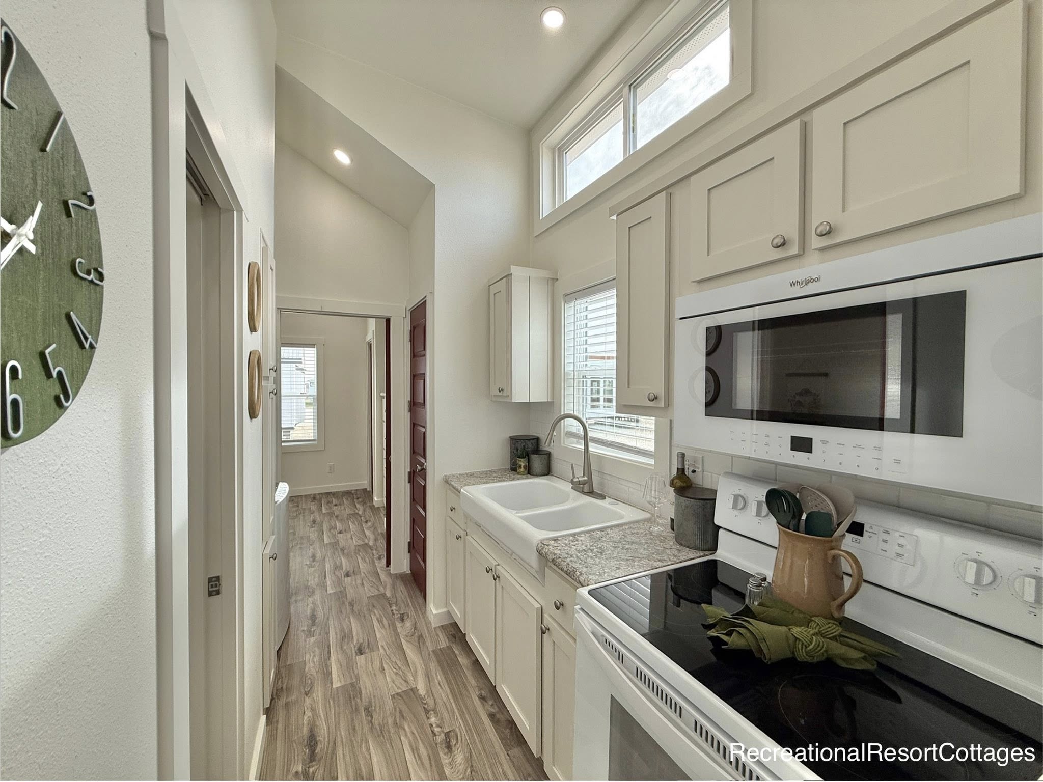 Compact modern farmhouse tiny home galley kitchen hallway with high vaulted ceilings, bright white shaker cabinets, farmhouse apron sink, gray quartz countertop, Whirlpool stainless steel appliances including gas range and microwave hood, spacious upper cabinets, clerestory windows for natural light, gray wood-look plank flooring, open layout leading to living areas and bedrooms – luxurious recreational resort cottage interior design perfect for tiny house, vacation cabin, and small space minimalist living inspiration.