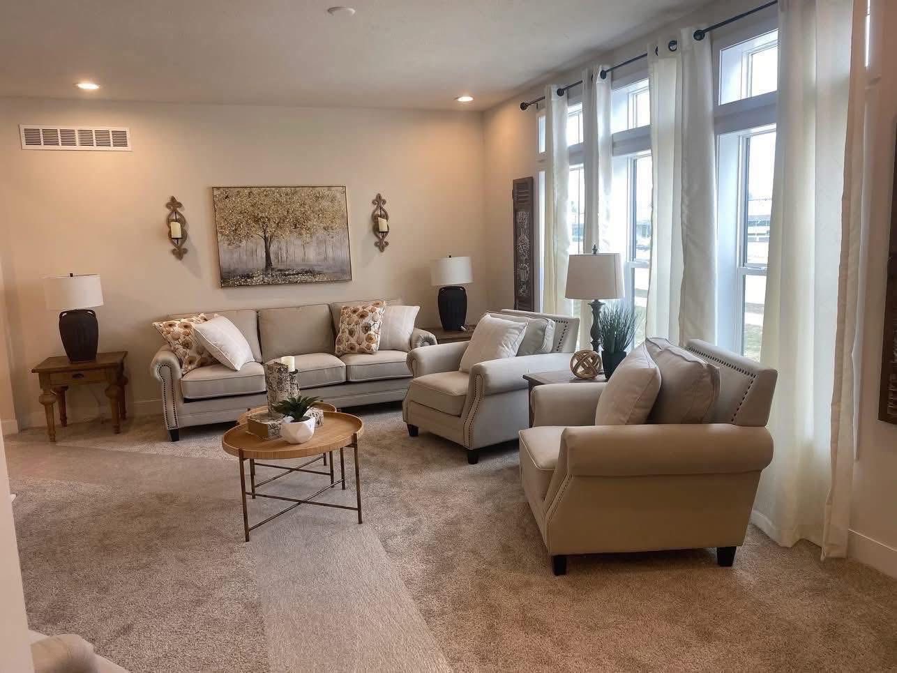 Luxurious spacious living room interior of a modern 3-bedroom customizable modular home featuring neutral beige tones, plush sectional sofa and armchair seating, elegant throw pillows, cozy area rug, large windows with sheer white curtains for natural light, contemporary tree artwork, decorative wall sconces, wooden side tables, potted plants, and warm ambient lighting creating an inviting family-friendly open-concept living space in The Garnet giveaway home.