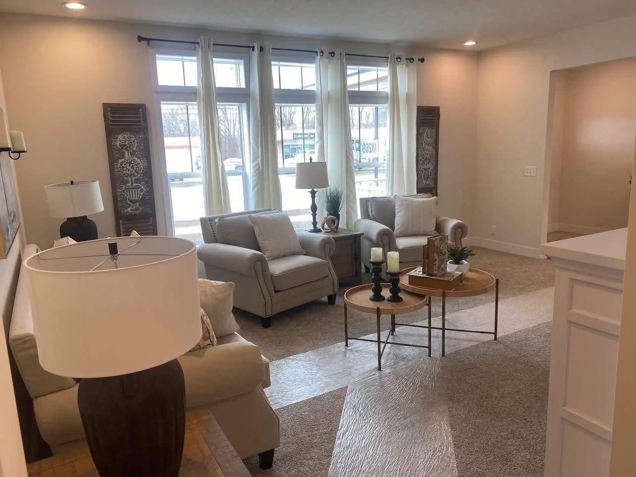 Elegant and inviting open-concept living room in a modern 3-bedroom customizable modular home featuring neutral beige and warm taupe tones, plush tufted sofa and matching armchairs with decorative throw pillows, large floor-to-ceiling windows draped in sheer white curtains flooding the space with natural light, textured area rug over hardwood-look flooring, rustic wooden accent tables with potted greenery and candles, tall carved wooden decorative panels, contemporary tree artwork, recessed ceiling lights, cozy family seating arrangement, and seamless flow to adjacent kitchen area creating a bright spacious family-friendly living space in The Garnet dream home giveaway.