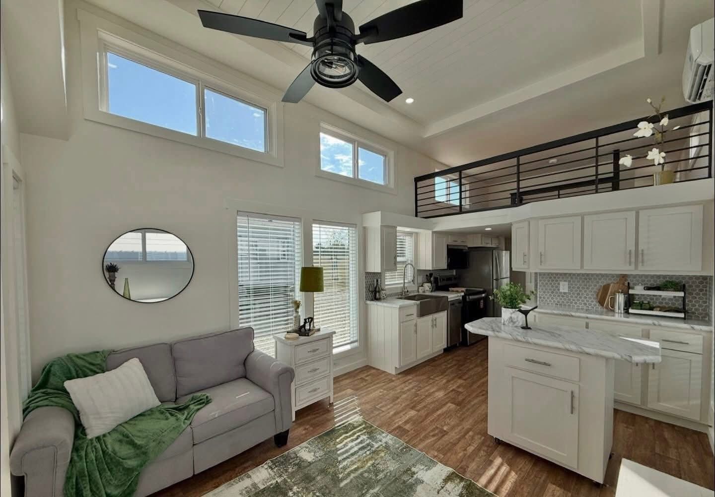 Modern living room with high ceilings, featuring a gray couch, green accents, and a small kitchen with white cabinets. Bright and welcoming atmosphere.