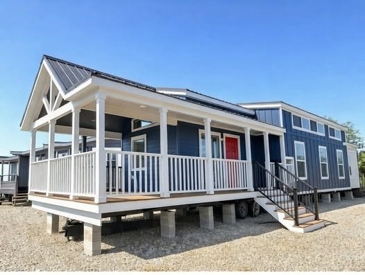 A modern blue tiny house with a white railing porch and bright red door, elevated on concrete blocks, sits under a clear blue sky, conveying warmth.