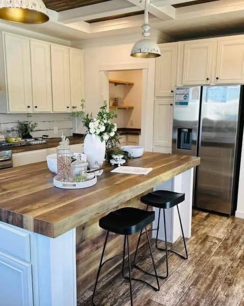 Elegant modern farmhouse kitchen in a customizable modular home featuring timeless cream shaker cabinets with black knob hardware, stunning reclaimed wood-look butcher block island countertop with rich natural grain and live-edge details seating two sleek black metal bar stools, spacious layout with abundant natural light from large windows and open arched doorway to adjacent rooms, stainless steel double-door refrigerator with water dispenser, white subway tile backsplash, gold-toned industrial pendant lights and vaulted ceiling with exposed dark wooden beams, decorative white vase with fresh greenery and flowers on island, glass canisters of pantry staples, woven tray accents, and cozy inviting family gathering space perfect for cooking entertaining and daily living in this luxurious dream manufactured home design.