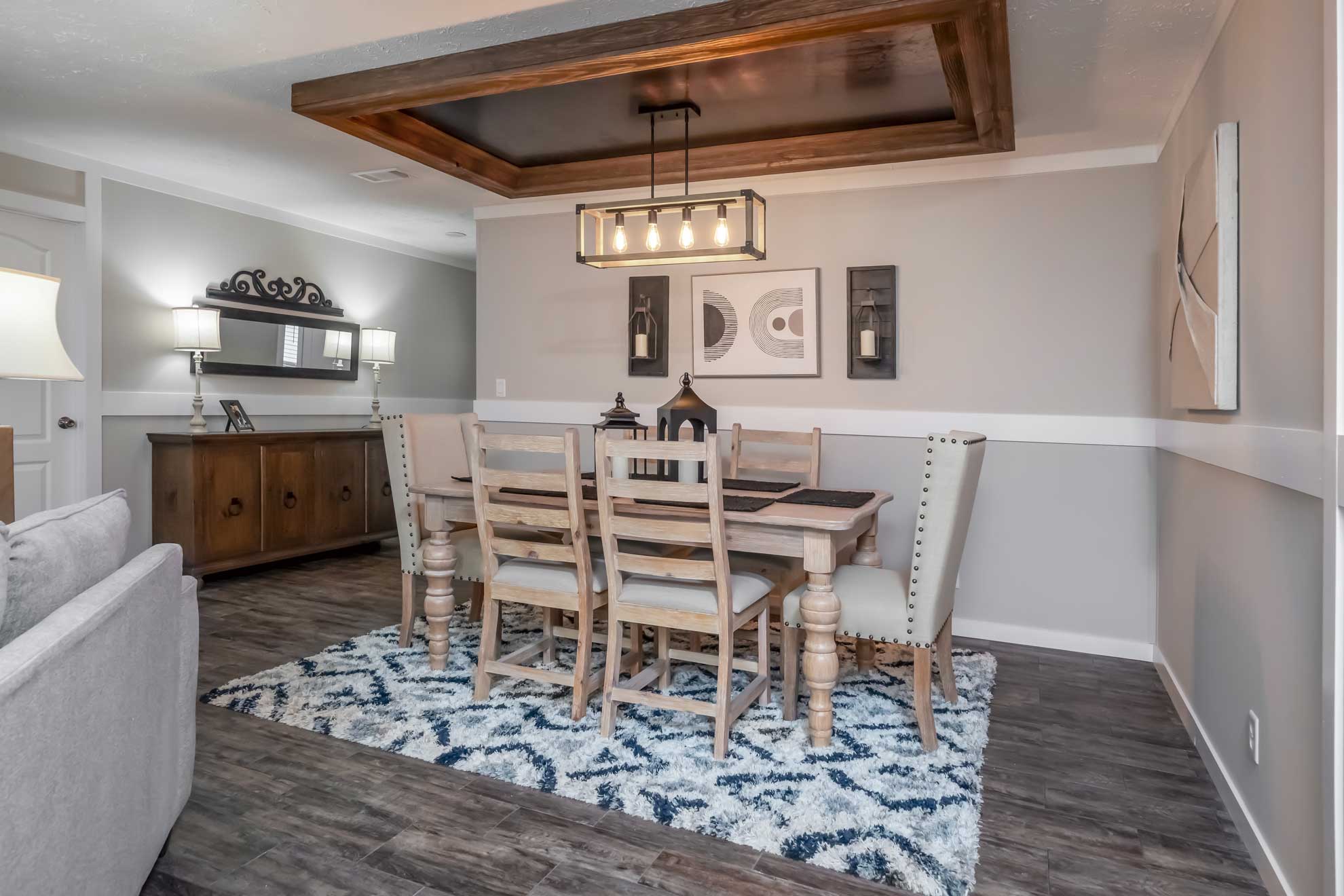 SEO-optimized alt text for a modern manufactured home dining room featuring a rustic wooden table set for six with upholstered and ladder-back chairs, positioned over a blue and white geometric area rug; the sophisticated interior design showcases a unique recessed tray ceiling with a wood-beam frame and a contemporary black metal linear chandelier, complemented by a dark wood sideboard and elegant wall decor to highlight premium mobile home floor plans and stylish modular home dining area ideas for maximum search engine visibility and ranking.