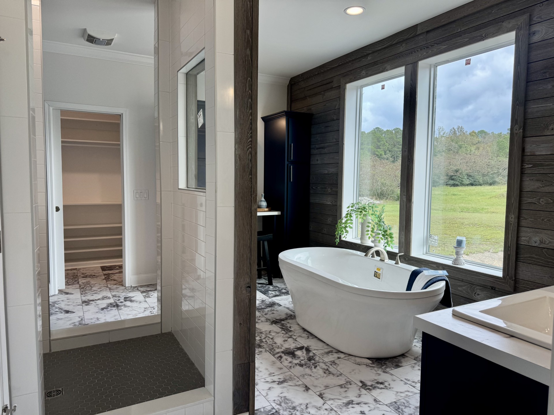 SEO-optimized alt text for a high-ranking luxury custom modular home master bathroom featuring a professional rustic-modern interior design with a deep white freestanding soaking tub and a spacious walk-in shower with charcoal hexagonal floor tiles; the architectural view showcases elegant marble-patterned porcelain floor tile, a warm wood-plank accent wall, and large double windows overlooking a serene landscape, complemented by a sophisticated navy blue vanity and a glimpse into a large walk-in closet with custom shelving, perfectly highlighting luxury prefab home floor plans, modern farmhouse bathroom decor ideas, and high-end residential mobile living finishes for maximum search engine visibility and ranking. Would you like me to create a description for the matching master bedroom or the modern kitchen next?