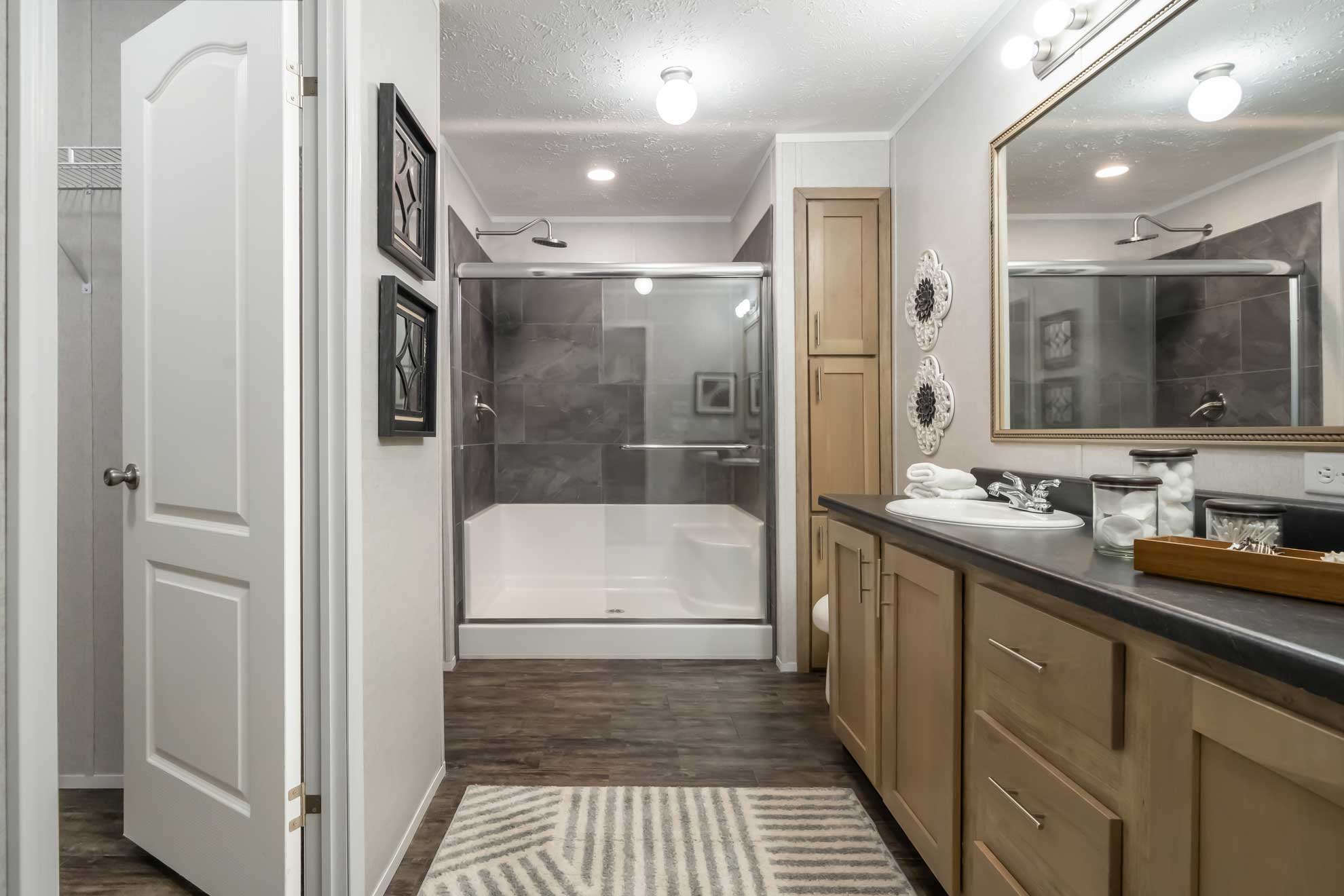 SEO-optimized alt text for a modern master bathroom in a luxury manufactured home featuring a spacious walk-in shower with gray stone-patterned tile, a built-in seat, and sliding glass doors; the high-ranking interior design includes a large vanity with light oak-finished cabinetry, a dark countertop with an integrated sink, and a large framed mirror, all complemented by durable wood-plank flooring and stylish decor that highlights premium mobile home bathroom renovations and contemporary modular home floor plans for maximum search engine visibility and ranking.