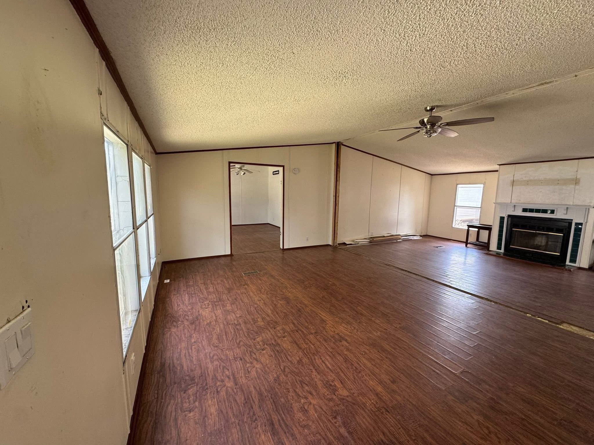 SEO-optimized alt text for an open-concept living room in an older manufactured home featuring expansive wood-look laminate flooring and a white brick fireplace with a black insert; the high-ranking interior view showcases a spacious layout with vaulted ceilings, a central ceiling fan, and large windows that provide natural light, perfectly highlighting affordable mobile home floor plans and renovation potential for maximum search engine visibility and ranking.