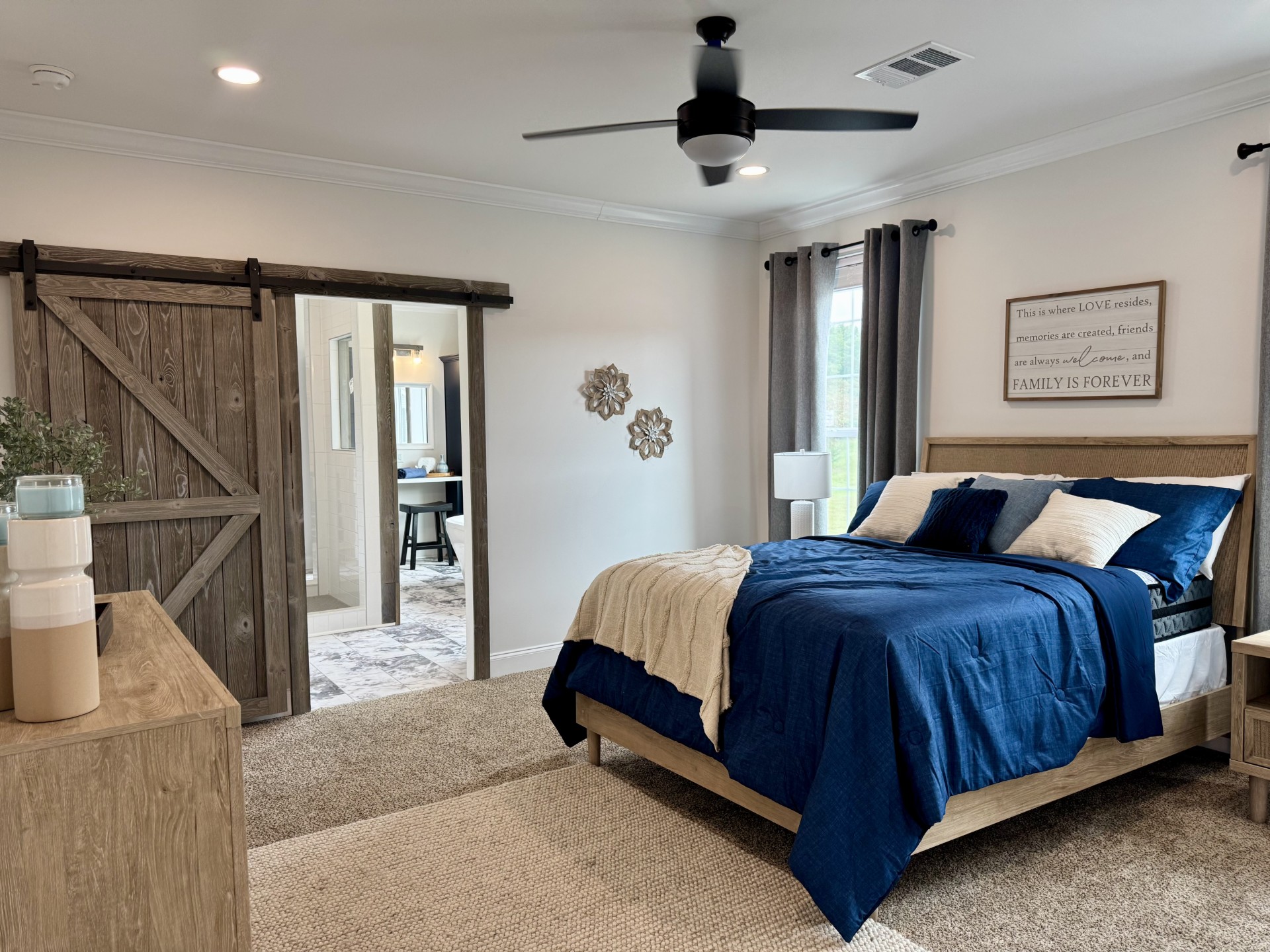 SEO-optimized alt text for a high-ranking luxury custom modular home master bedroom featuring a professional rustic-modern interior design with a sliding wood barn door leading to an en-suite bathroom; the architectural view showcases a cozy queen-sized bed with navy blue linens and a beige throw, a contemporary light-wood dresser, and soft neutral carpeting, complemented by a minimalist "Family Is Forever" wall art, a black ceiling fan, and elegant gray drapes over a sunlit window, perfectly highlighting luxury prefab home floor plans, modern farmhouse bedroom decor ideas, and high-end residential mobile living finishes for maximum search engine visibility and ranking.