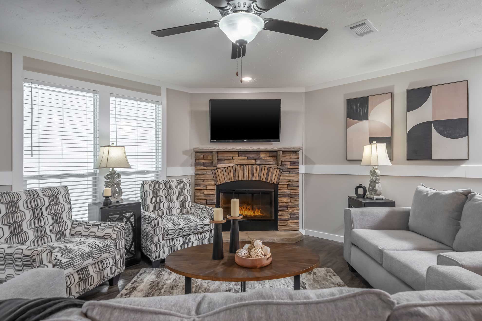 SEO-optimized alt text for a modern living room in a luxury manufactured home featuring a corner stone fireplace with a rustic wood mantel, a wall-mounted TV, and high-end furnishings including patterned accent chairs and a gray sectional sofa; the sophisticated interior design showcases light gray walls with white decorative trim, large windows with blinds for natural light, and contemporary decor elements that highlight premium mobile home floor plans and stylish modular home living area ideas for maximum search engine visibility and ranking.
