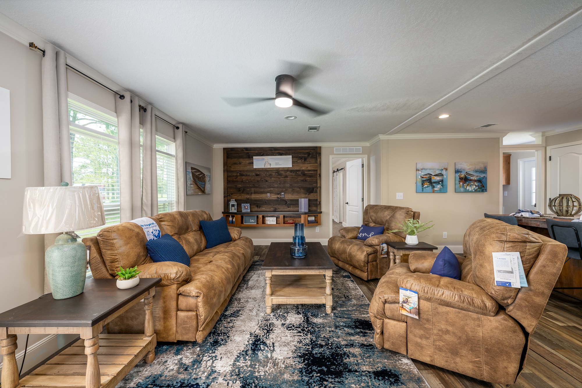 Cozy and spacious open-concept living room in The Garnet 1,873 sq ft 3-bedroom customizable modular home featuring warm neutral taupe walls, plush brown leather sectional sofa and matching recliners with navy blue accent pillows, large rustic wooden coffee table and side tables, textured blue-gray area rug over hardwood-look flooring, ceiling fan with integrated light, built-in wooden accent wall with open shelving displaying nautical decor lanterns and plants, coastal-themed boat artwork, sheer gray curtains on large windows flooding the space with natural light, potted greenery, and seamless flow to adjacent kitchen creating an inviting comfortable family gathering and relaxation area in this dream home giveaway winner.
