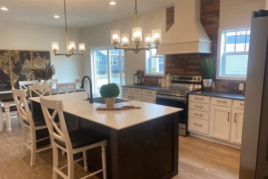 SEO-optimized alt text for a high-ranking luxury multi-section manufactured home kitchen and dining area featuring a professional open-concept interior design with a large dark-wood central island and white farmhouse-style barstools; the bright floor plan showcases a gourmet kitchen with premium white shaker cabinetry, a custom white range hood over a rustic wood-plank backsplash, and sophisticated gold-accented chandeliers, while the adjacent dining space features a matching dark-wood table and a three-panel vintage world map wall art to highlight spacious double-wide mobile home floor plans, upscale modular home decor, and luxury residential prefab home finishes for maximum search engine visibility and ranking.