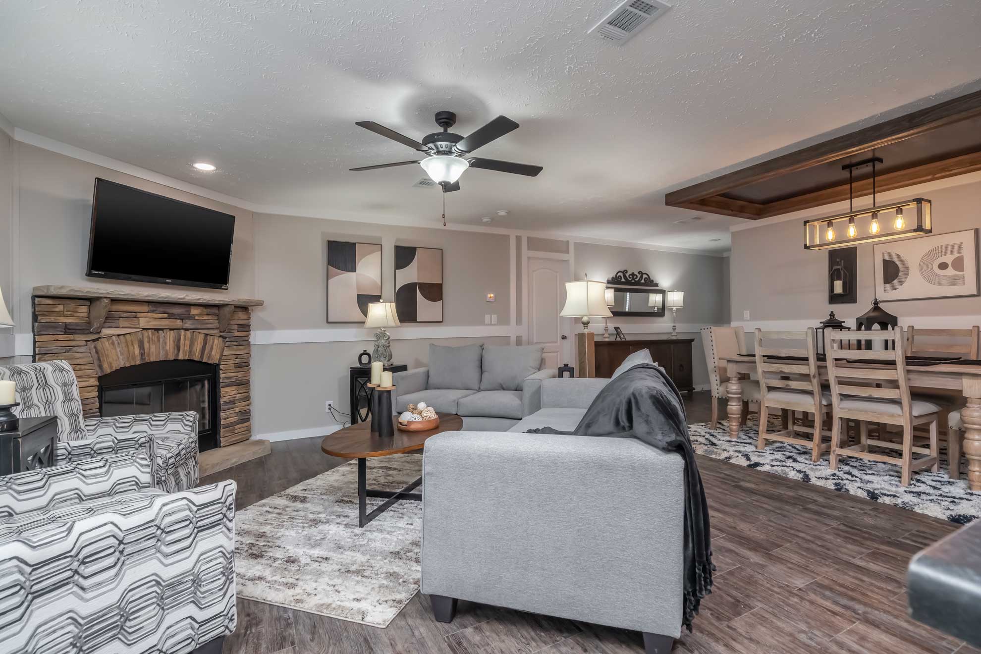 SEO-optimized alt text for a modern living room in a luxury manufactured home featuring a corner stone fireplace with a rustic wood mantel, a wall-mounted TV, and high-end furnishings including patterned accent chairs and a gray sectional sofa; the sophisticated interior design showcases light gray walls with white decorative trim, large windows with blinds for natural light, and contemporary decor elements that highlight premium mobile home floor plans and stylish modular home living area ideas for maximum search engine visibility and ranking.