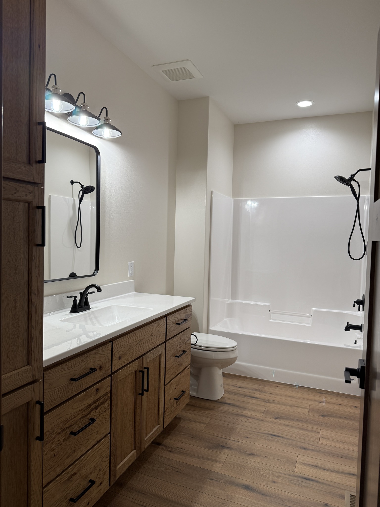 SEO-optimized alt text for a high-ranking modern luxury modular home bathroom featuring a professional double vanity with high-quality oak cabinetry and sleek white quartz countertops; the architectural interior design showcases a clean, minimalist aesthetic with contemporary black faucets, a large black-framed rectangular mirror, and a bright white alcove bathtub and shower combination, all set against durable wide-plank wood flooring and illuminated by a stylish three-light vanity fixture with Edison bulbs, perfectly highlighting premium prefab home floor plans, upscale craftsman-style bathroom finishes, and sophisticated residential mobile living solutions for maximum search engine visibility and ranking.