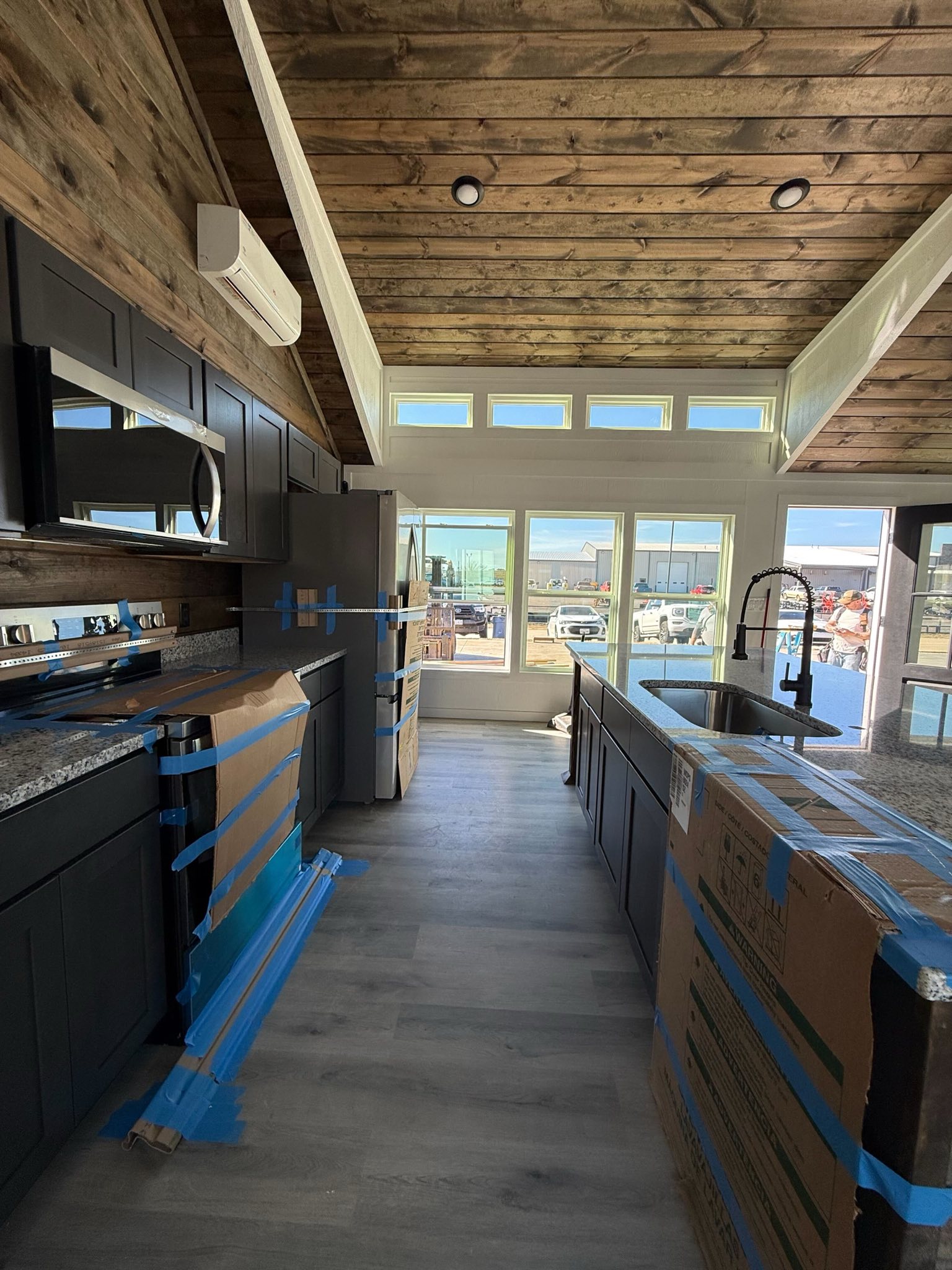 SEO-optimized alt text for a high-ranking luxury custom park model tiny home interior under construction, featuring a professional modern kitchen and open-concept living area; the architectural view showcases a high-end vaulted wood-plank ceiling with integrated recessed lighting, sophisticated matte black cabinetry with granite countertops, and a large central kitchen island with a deep stainless steel sink and industrial gooseneck faucet, all illuminated by a row of elegant transom windows and expansive sunlit glass doors to highlight premium small-space floor plans, upscale prefab tiny house interior finishes, and luxury residential mobile home construction for maximum search engine visibility and ranking.

Would you like me to provide the alt text for the matching master bedroom with the wood-plank accent wall next?