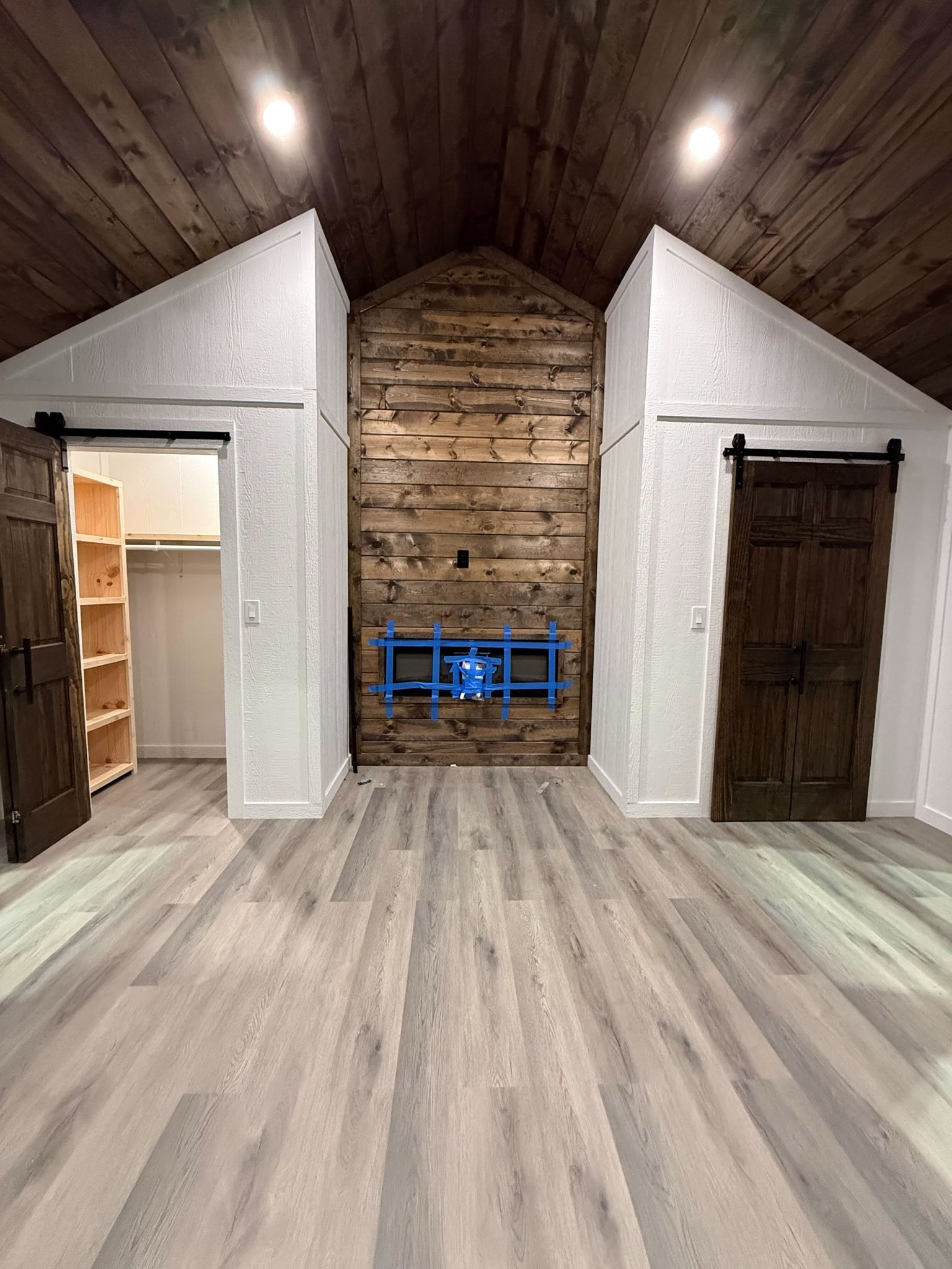 SEO-optimized alt text for a high-ranking luxury custom park model tiny home interior featuring a professional modern master suite design with a stunning vaulted dark-wood ceiling and premium wide-plank flooring; the architectural view showcases a central floor-to-ceiling rustic wood-plank accent wall with a built-in electric fireplace mounting area, flanked by two elegant dark-wood sliding barn doors leading to a spacious walk-in closet with custom shelving and an en-suite bathroom, perfectly highlighting luxury prefab tiny house floor plans, upscale small-space bedroom decor, and high-end residential mobile living solutions for maximum search engine visibility and ranking.