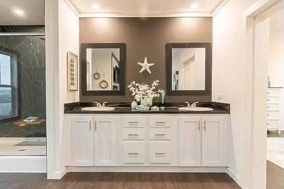 SEO-optimized alt text for a high-ranking luxury multi-section manufactured home master bathroom featuring a professional modern spa-like interior design with a large white double vanity; the bright, spacious layout showcases premium white shaker-style cabinetry with gold hardware, a sophisticated dark stone countertop with dual undermount sinks, and two large dark-framed mirrors set against a contemporary taupe accent wall, all complemented by a glimpse of a walk-in glass shower with custom marble-patterned tile to highlight spacious double-wide mobile home floor plans, upscale modular bathroom decor, and luxury residential prefab home finishes for maximum search engine visibility and ranking.