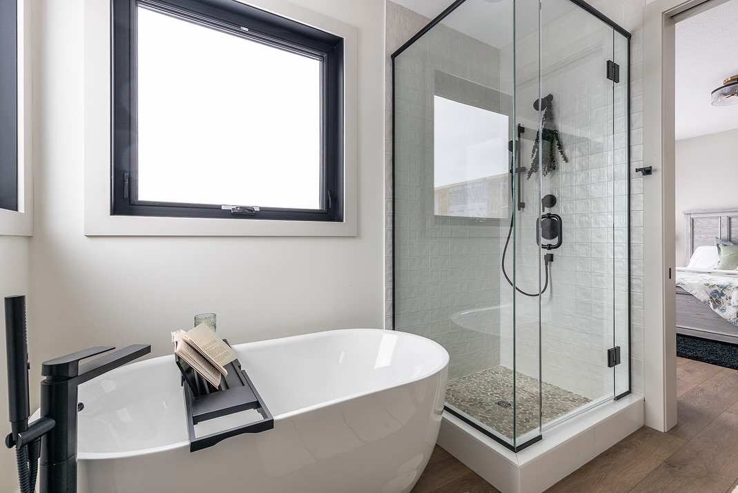 SEO-optimized alt text for a high-ranking luxury custom modular home master bathroom featuring a professional modern interior design with a sleek white freestanding soaking tub and a contemporary glass-enclosed walk-in shower; the architectural view showcases premium black matte fixtures, a stylish black-framed window for natural light, and elegant white vertical subway tile, perfectly highlighting luxury prefab home floor plans, upscale craftsman-style bathroom finishes, and high-end residential mobile living solutions for maximum search engine visibility and ranking.