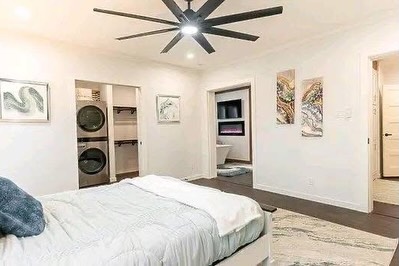 SEO-optimized alt text for a high-ranking luxury multi-section manufactured home master suite featuring a professional modern interior design with a sophisticated white and dark-gray color palette; the bright, spacious bedroom showcases a premium king-sized bed with plush white bedding, a contemporary large-scale black ceiling fan, and high-end wide-plank flooring, while the architectural layout reveals a functional laundry nook with a stacked front-load washer and dryer, a walk-in closet with custom shelving, and a glimpse into an upscale en-suite bathroom with a soaking tub and electric fireplace to highlight spacious double-wide mobile home floor plans, luxury modular bedroom decor, and premium residential prefab home finishes for maximum search engine visibility and ranking.