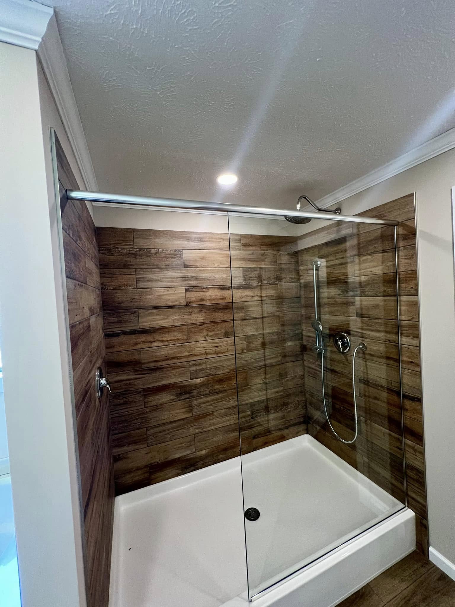 SEO-optimized alt text for a luxury manufactured home bathroom featuring a modern walk-in shower with rustic wood-plank tile walls, a sleek glass sliding door, and a clean white shower base; the high-end interior design showcases premium chrome fixtures including a rainfall showerhead and handheld sprayer, perfectly capturing contemporary small-space luxury and functional elegance for high-ranking searches related to modern mobile home renovations, sophisticated bathroom decor, and innovative modular home floor plans.