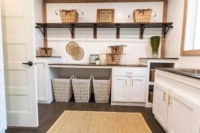 SEO-optimized alt text for a high-ranking luxury multi-section manufactured home utility and laundry room featuring a professional modern interior design with an expansive white cabinetry suite and warm wood-tone countertops; the bright, functional space showcases an integrated workstation with custom shelving, premium white shaker-style cabinets with gold hardware, and a dedicated laundry area with large woven sorting baskets and a built-in sink, all set against high-end wide-plank flooring and a natural jute area rug to highlight spacious double-wide mobile home floor plans, upscale modular laundry room decor, and luxury residential prefab home finishes for maximum search engine visibility and ranking.