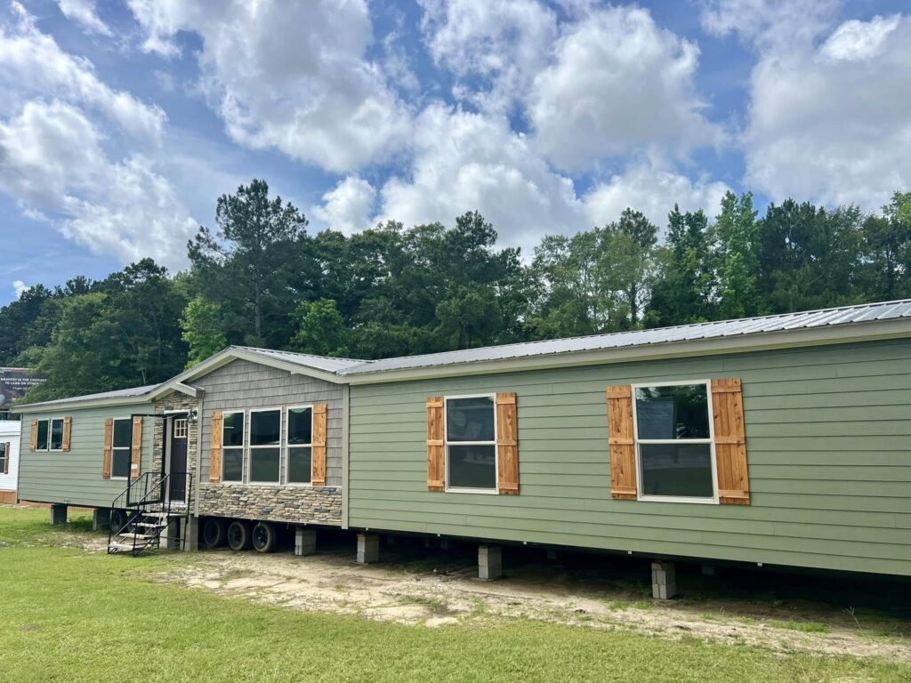 SEO-optimized exterior view of a large, high-end double-wide manufactured home featuring olive green horizontal siding, rustic cedar wood shutters, and a sophisticated stone-veneer entryway under a gable roof; the long-profile mobile home design includes a durable silver metal roof and multiple large energy-efficient windows, set on a gravel foundation against a backdrop of lush green trees and a bright cloudy sky, perfectly showcasing modern curb appeal for luxury modular housing and premium off-site residential construction.