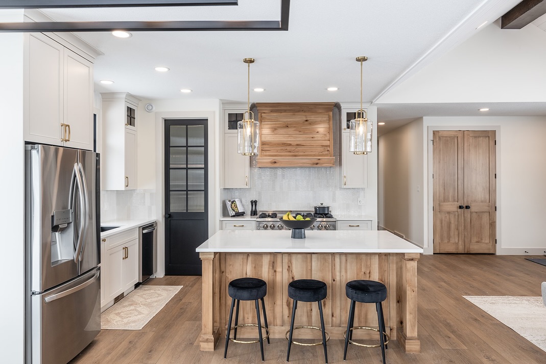 SEO-optimized alt text for a high-ranking luxury custom modular home kitchen featuring professional-grade white cabinetry and a large rustic wood kitchen island with a white quartz countertop; the sophisticated interior design showcases a premium wood-veneer range hood, elegant white vertical subway tile backsplash, and contemporary gold hardware, complemented by stylish black velvet barstools, modern glass pendant lighting, and a high-end stainless steel refrigerator, all set against durable wide-plank oak flooring to highlight gourmet prefab kitchen layouts, sophisticated open-concept floor plans, and upscale residential mobile living finishes for maximum search engine visibility and ranking.