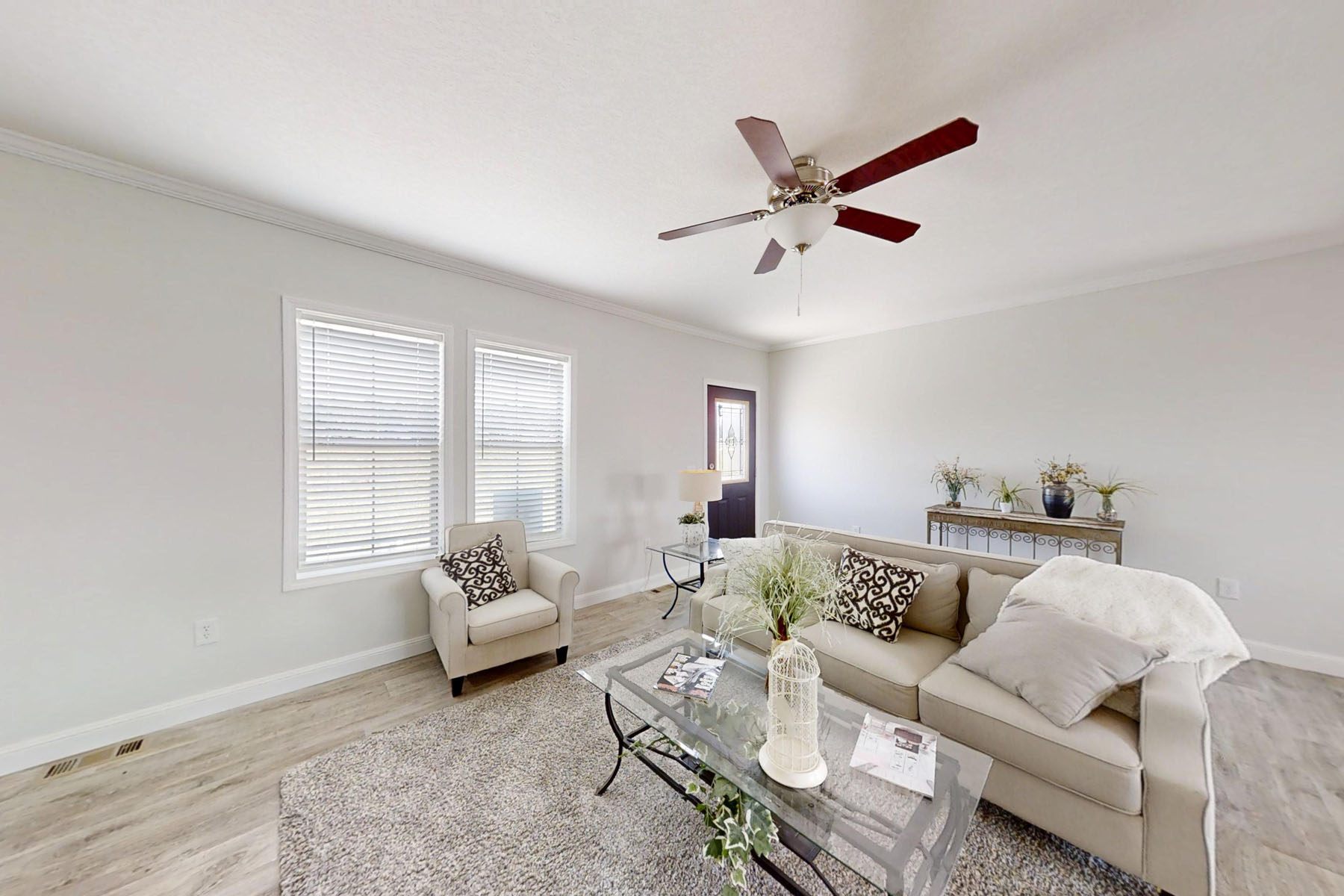 
Gemini said
Modern living room interior design featuring a neutral beige sofa with decorative patterned pillows, a matching armchair, and a glass-top coffee table on a plush grey area rug, complemented by light-colored hardwood flooring, large windows with white blinds, and a classic ceiling fan, creating a bright and airy transitional home atmosphere perfect for residential real estate listings and home staging inspiration.