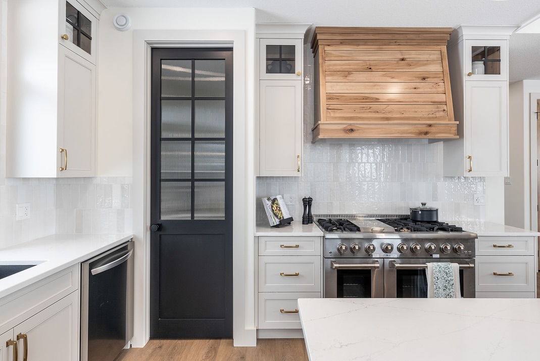 SEO-optimized alt text for a high-ranking luxury custom modular home kitchen featuring professional-grade white shaker cabinets and a premium stainless steel double-oven range; the sophisticated interior design showcases a rustic wood-veneer range hood, elegant white vertical subway tile backsplash, and contemporary gold hardware, complemented by a stylish black frosted-glass pantry door, white marble-style quartz countertops, and durable wide-plank oak flooring, perfectly highlighting gourmet prefab kitchen layouts, modern open-concept floor plans, and high-end residential mobile living finishes for maximum search engine visibility and ranking.