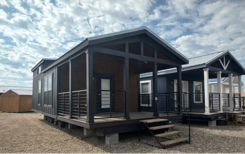 Modern tiny house with a dark wood facade, porch, and steps, set on gravel under a partly cloudy sky. Adjacent to similar structures.