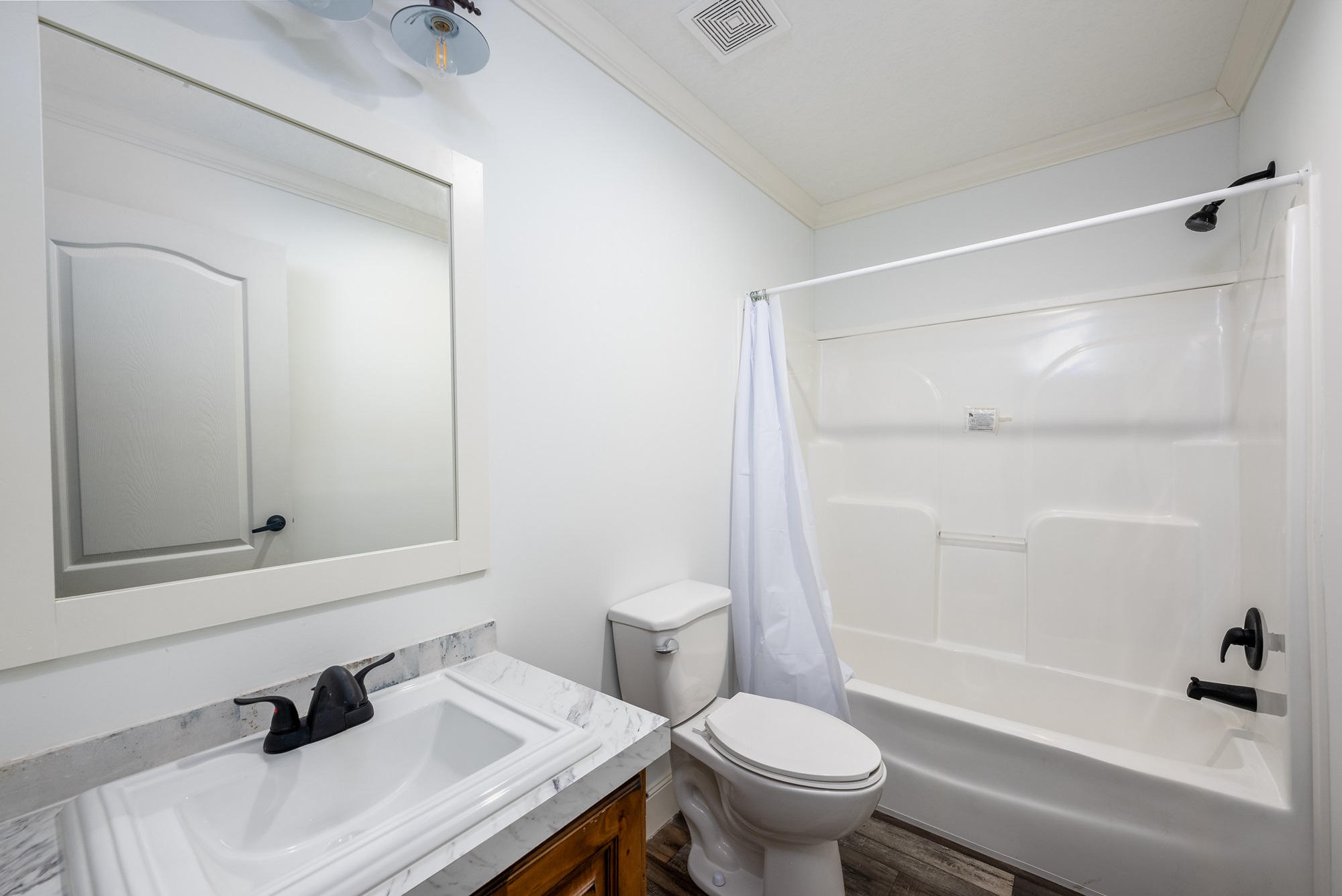 Compact functional secondary bathroom in The Garnet 1,873 sq ft 3-bedroom customizable modular home featuring clean white walls with crown molding, large rectangular vanity mirror with arched top frame over white marble-look quartz countertop and dark wood cabinet base with matte black faucet and hardware, standard white bathtub-shower combo with curved acrylic surround white curtain rod and liner, adjacent white toilet, textured ceiling with vent fan and Edison bulb vanity light fixture, warm wood-look flooring, bright airy layout with ample natural light creating a practical comfortable guest or family bathroom space in this affordable dream home giveaway winner.