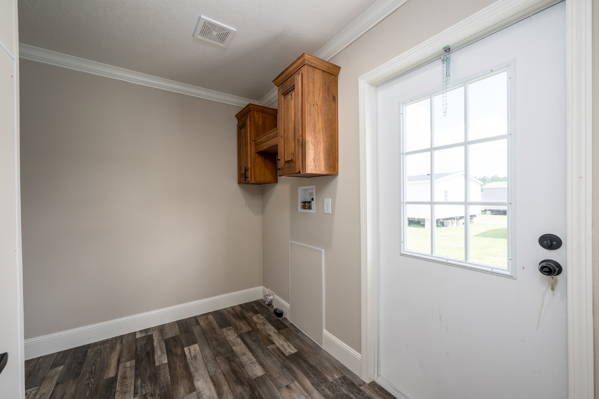 Spacious bright entryway mudroom laundry combo in The Garnet 1,873 sq ft 3-bedroom customizable modular home featuring neutral taupe walls with crisp white crown molding and baseboards, warm wood-look vinyl plank flooring, upper walnut wood cabinet storage with black hardware for organization, large glass-paneled front entry door with white frame and chain lock allowing abundant natural light, wall-mounted electrical panel and outlet, clean empty layout ready for washer dryer hookups bench hooks and personalization, creating a welcoming functional transition space from outdoors to family living areas in this affordable dream home giveaway winner.