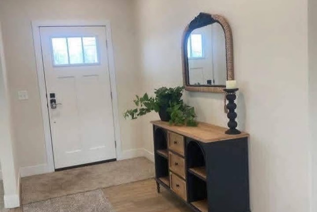 SEO-optimized alt text for a high-ranking luxury manufactured home entryway featuring a professional farmhouse interior design with a classic white craftsman-style front door; the bright foyer showcases a sophisticated black and natural wood console table decorated with lush green plants and a tall black candle holder, positioned beneath a stylish arched wicker-framed wall mirror to create a welcoming first impression, perfectly highlighting modern prefab home entry decor, upscale small-space furniture solutions, and premium residential modular home finishes for maximum search engine visibility and ranking.

Would you like me to generate SEO alt text for another interior or exterior view of this home?