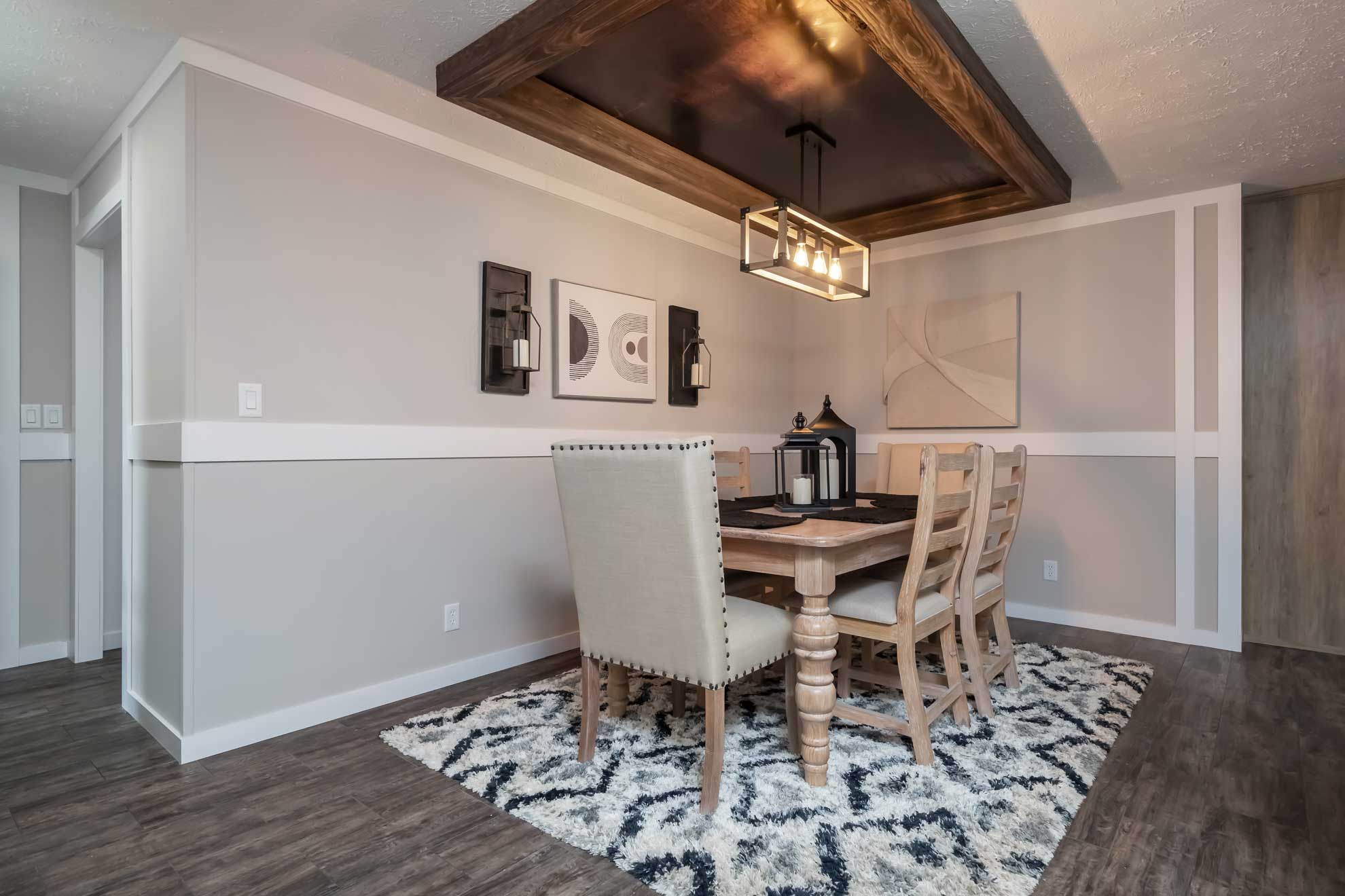SEO-optimized alt text for a modern manufactured home dining room featuring a rustic wooden table set with upholstered and ladder-back chairs, positioned over a blue and white geometric area rug; the sophisticated interior design showcases a unique recessed tray ceiling with a wood-beam frame and a contemporary black metal linear chandelier, complemented by a dark wood sideboard and elegant wall decor to highlight premium mobile home floor plans and stylish modular home dining area ideas for maximum search engine visibility and ranking.