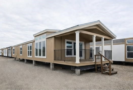 Row of modern farmhouse tiny homes on wheels in a manufactured housing community featuring tan board and batten siding with white trim, spacious covered front porches supported by elegant white columns, black metal railings, gabled metal roofs, large grid-pattern windows and transom glass for abundant natural light, elevated decks on trailer chassis with concrete block piers, wooden entry staircases with handrails, neutral earthy tones, overcast sky backdrop ideal for affordable modular living, mobile tiny house communities, off-grid setups, or vacation cabin developments.