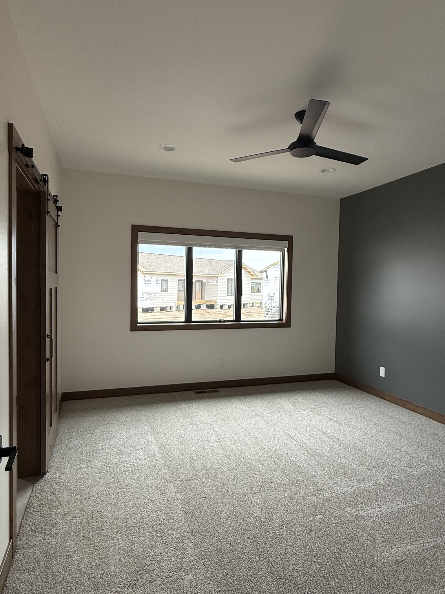 SEO-optimized alt text for a high-ranking modern luxury modular home bedroom featuring a professional interior design with a sophisticated dark gray accent wall and neutral beige carpeting; the spacious master suite showcases a contemporary black ceiling fan, large black-framed windows for natural light, and stylish sliding barn doors with dark wood trim, perfectly highlighting premium prefab home floor plans, trendy craftsman-style architectural details, and high-end residential mobile living solutions for maximum search engine visibility and ranking.