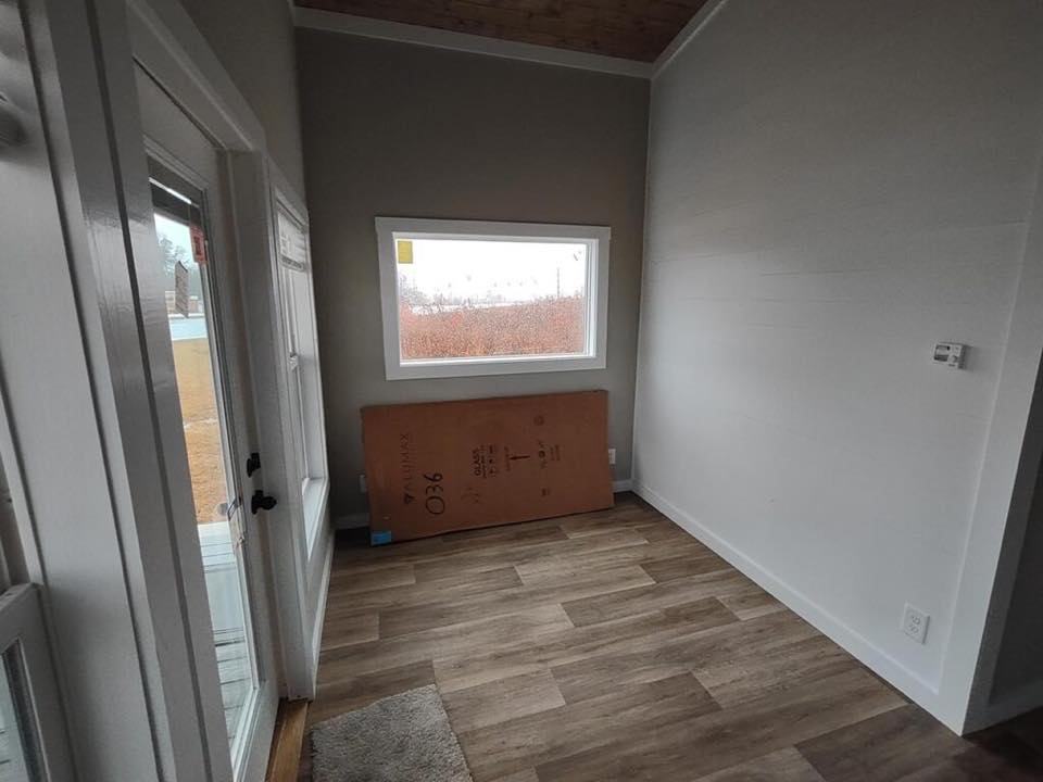 Small room with light gray walls, wooden floor, and a large window showing a cloudy exterior. A cardboard box leans against the wall, creating a minimalistic, unfinished feel.