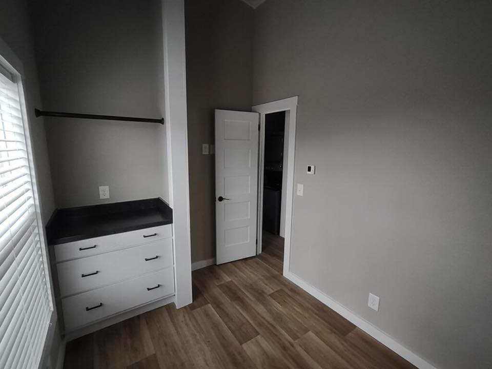 Minimalist room with light wood flooring, gray walls, and white trim. Features a small closet with drawers, black countertop, and an open white door.
