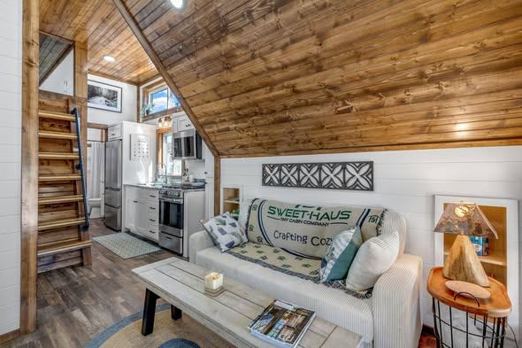 Cozy tiny house interior with wooden walls and ceiling. Features a white couch with green pillows, a small coffee table, a framed artwork, and a welcoming kitchen in the background.