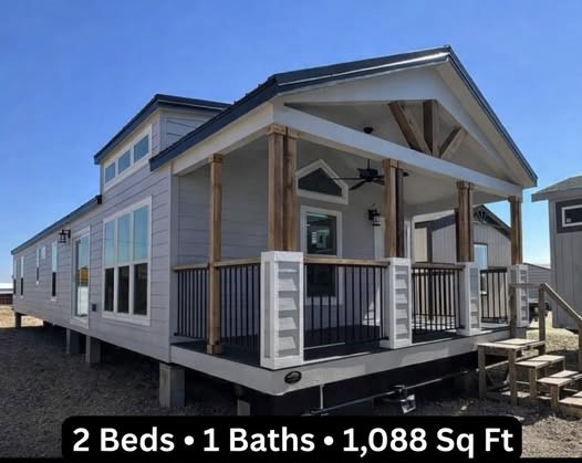 Modern luxury single-wide modular home featuring a light grey exterior with a spacious covered front porch, natural wood pillars, and black industrial-style railings. The 1,088 square foot floor plan includes 2 bedrooms and 1 bath, showcasing premium architectural details like a vaulted gable roof with decorative timber trusses, energy-efficient clerestory windows, and a sophisticated contemporary aesthetic perfect for high-end prefab or tiny home living.