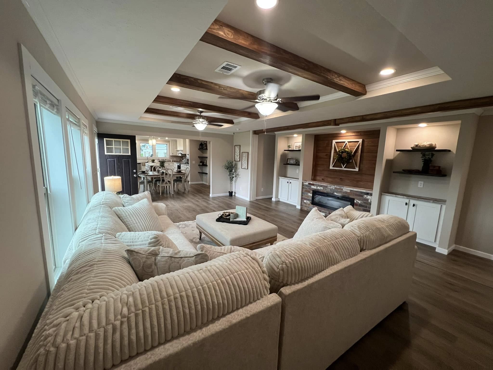 SEO-optimized alt text for a luxury manufactured home's open-concept living area, featuring a large cream-colored sectional sofa, a spacious tray ceiling with rustic wood beams and dual ceiling fans, and high-end wood-look flooring; the expansive interior design showcases a modern fireplace with a brick surround, built-in white cabinetry with dark floating shelves, and a clear view into a bright dining room and gourmet kitchen, perfectly highlighting premium mobile home floor plans, contemporary farmhouse decor, and sophisticated residential modular housing for maximum search engine visibility and ranking.