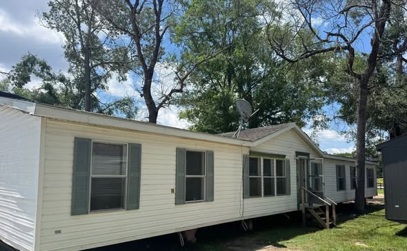 SEO-optimized alt text for an older double-wide manufactured home featuring cream-colored horizontal siding, forest green shutters, and a simple wooden porch entry; the high-ranking exterior view showcases a classic residential floor plan with a pitched roof, a satellite dish, and surrounding mature trees, perfectly highlighting affordable used mobile homes for sale and traditional modular housing options for maximum search engine visibility and ranking.