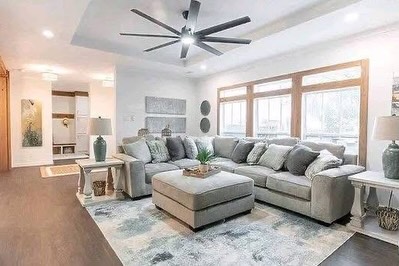 SEO-optimized alt text for a high-ranking luxury multi-section manufactured home living room featuring a professional modern interior design with a spacious gray sectional sofa and a large matching ottoman; the bright, open-concept floor plan showcases a contemporary tray ceiling with a high-end multi-blade ceiling fan, expansive wood-framed windows that provide abundant natural light, and a stylish abstract area rug over premium wide-plank flooring, perfectly highlighting spacious double-wide mobile home floor plans, upscale modular living room decor, and luxury residential prefab home finishes for maximum search engine visibility and ranking.