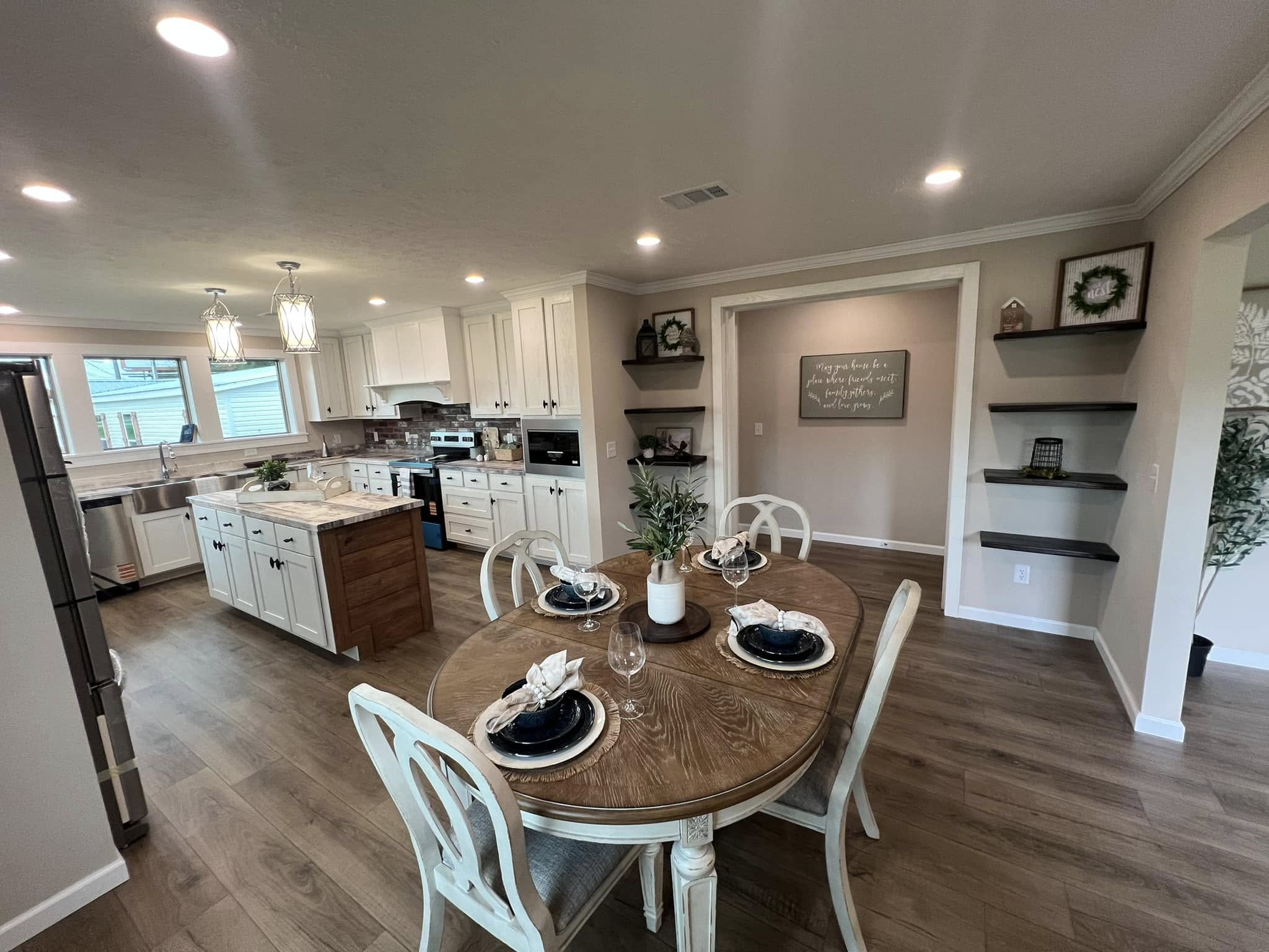 High-ranking SEO-optimized alt text for a luxury open-concept manufactured home interior, featuring a gourmet kitchen with white shaker cabinets, a rustic wood-top island, and red brick backsplash alongside a sophisticated dining area with a round wooden table and elegant chairs; the expansive floor plan showcases premium wood-look flooring, modern pendant lighting, and a bright living room with a tray ceiling and exposed wood beams, perfectly highlighting contemporary farmhouse decor and high-end modular home design for maximum search visibility.