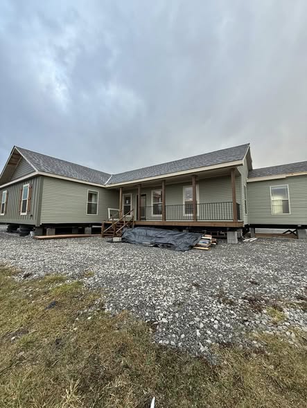 Modern farmhouse modular home exterior under dramatic cloudy sky featuring sage green vertical board and batten siding, spacious wraparound covered porch with wooden columns and black metal railings, gabled roof with gray asphalt shingles, large energy-efficient windows with white frames, elevated foundation on concrete piers with gravel base, wooden front steps leading to double entry doors, rustic natural wood porch accents, contemporary rural prefab tiny house or manufactured cottage design ideal for countryside living, vacation retreat, or off-grid family residence with timeless curb appeal